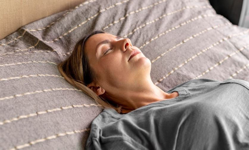 Mujer descansando pacíficamente en la cama con ropa casual, sobre una colcha gris con líneas decorativas, en un ambiente tranquilo y cómodo.