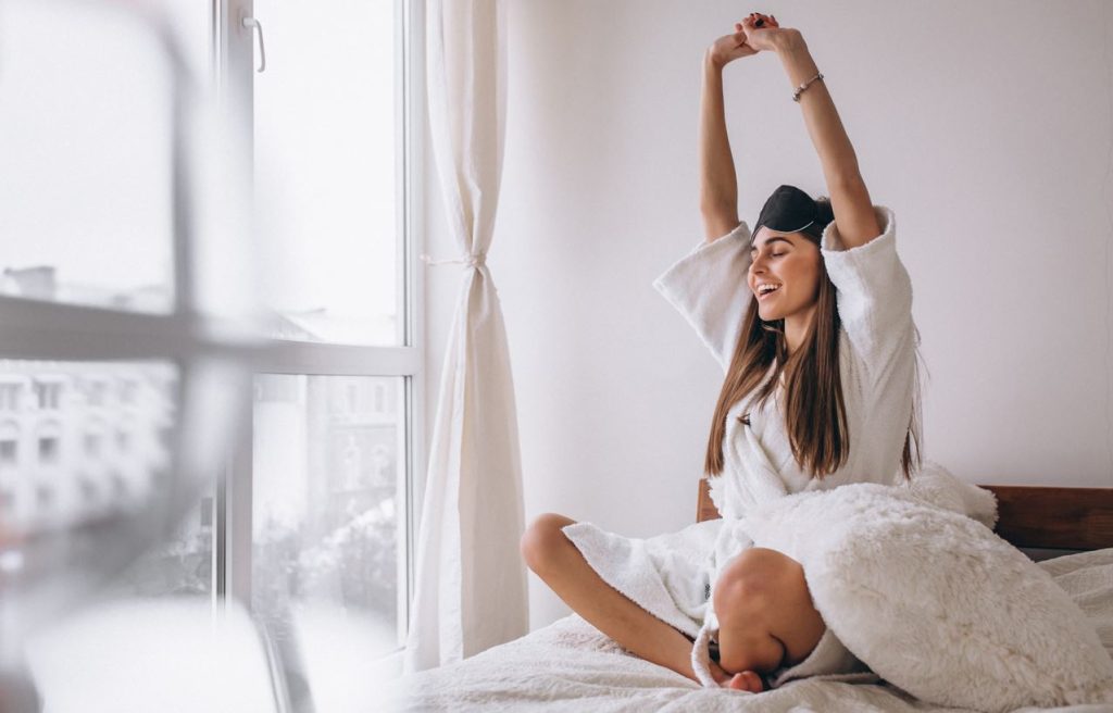 Mujer sonriente estirándose en la cama después de despertar, con una antifaz de dormir levantado sobre su frente, rodeada de luz natural y sábanas blancas, transmitiendo una sensación de energía matutina y bienestar.