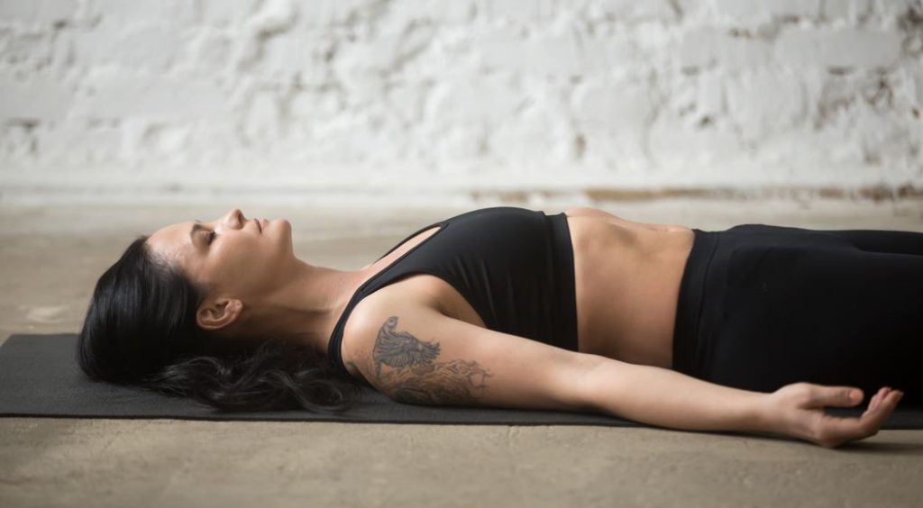 Mujer practicando Yoga Nidra o NSDR en una esterilla, vestida con ropa deportiva negra y luciendo un tatuaje de águila en el brazo, en un espacio de meditación minimalista con pared de ladrillo blanco.