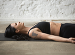 Mujer practicando Yoga Nidra en una esterilla, vestida con ropa deportiva negra y luciendo un tatuaje de águila en el brazo, en un espacio de meditación minimalista con pared de ladrillo blanco.